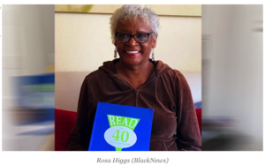 Older African American woman with black rimmed glasses holding a blue book. She has on a dark brown sweater and has gray hair. She is smiling