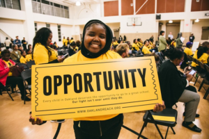 Girl holding big yellow sign that says "Opportunity". She is also wearing a yellow shirt. Behind here is a crowd in what looks like a school auditorium.